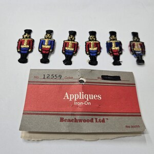 May include: Six embroidered iron-on appliques of toy soldiers in red, blue, and gold uniforms. The appliques are arranged above a red and gray tag that reads "Appliques Iron-On" and "Beachwood Ltd".