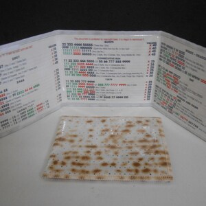 May include: A folded, clear plastic card with printed text and a rectangular, flat, unleavened bread. The card is open, displaying lists of numbers and words. The bread is light brown with darker brown spots.