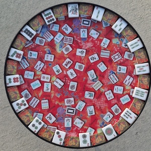 May include: Round table with a red and orange patterned surface, scattered with white Mahjong tiles. The table has a black rim and a decorative border with butterfly and floral designs. A classic game table.
