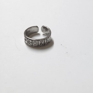 May include: A silver-colored adjustable ring with engraved Chinese characters. The ring has a slightly oxidized finish and is open at the top. The ring is on a white surface.