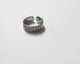 May include: A silver-colored adjustable ring with engraved Chinese characters. The ring has a slightly oxidized finish and is open at the top. The ring is on a white surface.