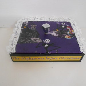 May include: A decorative box featuring imagery from "The Nightmare Before Christmas." The box has a purple top with a printed design and a white lace trim. The base is black with yellow text that reads "The Nightmare Before Christmas."