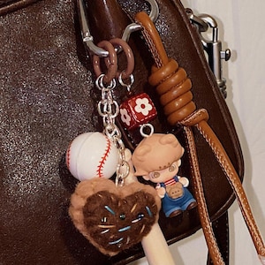 May include: A brown leather purse with a silver clasp and a collection of keychains. The keychains include a baseball, a heart-shaped charm, a small figure, and a red and white floral cube.