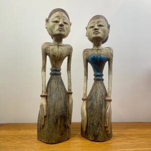May include: Two wooden statues of people, painted in shades of white, blue, and brown. The statues are standing upright with their hands clasped in front of them.