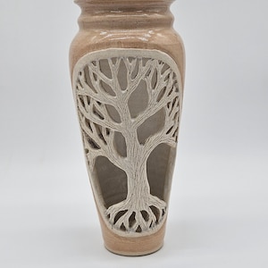 May include: A tan ceramic vase with a tree design carved into the front. The tree is white and has detailed branches and roots. The vase has a slightly textured surface and a wide opening at the top.
