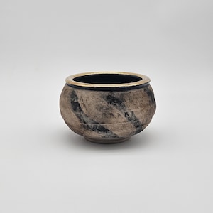 May include: A small, round ceramic bowl with a textured, earthy appearance. The bowl features a dark rim and a gold-colored interior rim. The exterior has a mottled pattern of brown and black, creating a rustic aesthetic. The bowl is set against a plain white background.