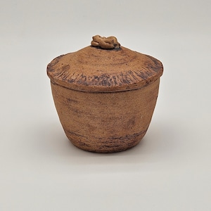 May include: A handmade, brown ceramic jar with a lid. The jar has a rounded shape with a textured surface and a small, decorative knob on the lid. The color is a warm, earthy brown, suggesting a rustic or artisanal style.