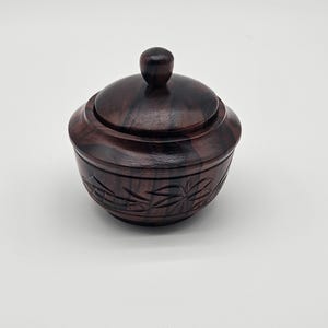 May include: A small, round, dark brown wooden bowl with a matching lid featuring a knob handle. The exterior of the bowl displays a natural wood grain pattern with darker streaks and a carved design of leaves encircling the lower section.