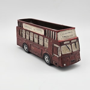 May include: A decorative, red double-decker bus planter with white accents. The bus has the text "CHAMPAGNE DE COGNAC" and "WE'RE GOING TO TIMBER CITY" on the top. The planter has a rectangular opening for plants.
