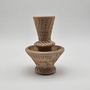 May include: A light brown, unglazed clay vessel featuring a wide, bowl-shaped base and a narrower, cylindrical top section. The entire surface is decorated with intricate, hand-carved geometric patterns, including small indentations and lines.