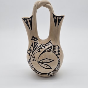 May include: A beige-colored, handcrafted pottery vase with a unique handle. The vase features intricate black geometric designs and patterns. The handle is a twisted loop, adding to its artistic appeal. This decorative piece is ideal for home decor.