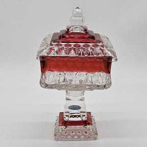 May include: A clear and red glass candy dish with a lid. The dish has a square base and a pedestal. The lid is also square and has a clear glass finial. The dish is likely made of crystal.