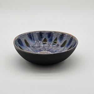 May include: A handmade ceramic bowl with a black exterior and a blue and green interior. The bowl has a textured, ridged design on the inside, creating a unique visual effect. The rim of the bowl has a golden-brown color.