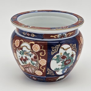 May include: A round, ceramic planter with a wide opening. The planter features a dark red and navy blue background with floral and bird designs. Gold trim accents the rim and the designs. The interior is white.