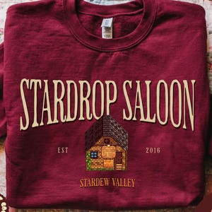May include: A garnet-colored sweatshirt featuring the text "STARDROP SALOON" in a vintage font. Below the text is an illustration of a saloon with the text "EST 2016" and "STARDEW VALLEY". The sweatshirt has a crew neck and long sleeves.