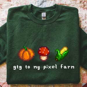 May include: Forest green sweatshirt with pixelated graphics of a pumpkin, mushroom, and corn. The text "gtg to my pixel farm" is printed below the graphics. The sweatshirt is made of a soft material.