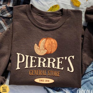 May include: A chocolate-colored sweatshirt with the text "Pierre's General Store" and a pumpkin graphic. The sweatshirt also features the text "Since 2016". The words "Witty Print Goods" are also visible.