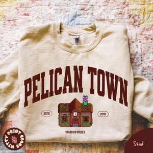 May include: Sand-colored sweatshirt with the text "PELICAN TOWN" in maroon arched lettering. Below the text is a graphic of a building with a clock, and the words "ESTD" and "2016" on either side. The words "STARDEW VALLEY" are below the building graphic.