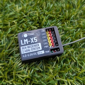 以下が含まれることがあります： 黒いLM-X5 5CHレシーバー2.4GHzシステム。カラフルなピンと「4.0-6.0V/DC」の文字があります。緑の草の上に置かれています。