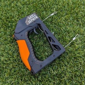 Puede incluir: Herramienta Line Master Zephyr negra y naranja con gatillo y dos cables met&aacute;licos. La herramienta tiene un mango con agarre naranja y el texto "Line Master ZEPHYR" en la parte superior. La herramienta est&aacute; sobre hierba verde.