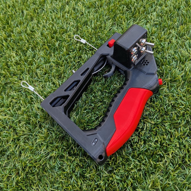 Puede incluir: Un dispositivo port&aacute;til negro y rojo con gatillo, interruptores y empu&ntilde;adura. El dispositivo tiene un marco rectangular con dos bucles met&aacute;licos adjuntos. La empu&ntilde;adura es roja y el resto del dispositivo es negro.