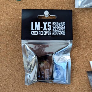 Może przedstawiać: Odbiornik LM-X5 5CH w srebrnym, szczelnie zamkniętym plastikowym opakowaniu z czarną kartą nagł&oacute;wkową. Nagł&oacute;wek zawiera tekst LM-X5 5CH RECEIVER oraz kod QR. Odbiornik wewnątrz jest oznaczony jako LM-X5 5CH RECEIVER 2.4GHz SYSTEM z widocznymi portami.