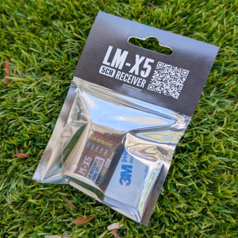 Może przedstawiać: Odbiornik LM-X5 5CH w przezroczystej plastikowej torebce z czarną kartą nagł&oacute;wkową. Karta zawiera napis LM-X5 5CH RECEIVER oraz kod QR. Odbiornik wewnątrz jest oznaczony jako LM-X5 CH RECEIVER 2.4GHz SYSTEM 4.0-6.0V DC i zawiera pasek samoprzylepny 3M.