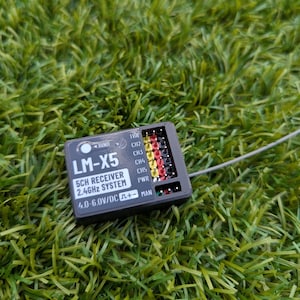 Puede incluir: Un receptor LM-X5 5CH negro, sistema de 2,4 GHz, con pines de colores y el texto "4.0-6.0V/DC". El dispositivo est&aacute; sobre hierba verde.
