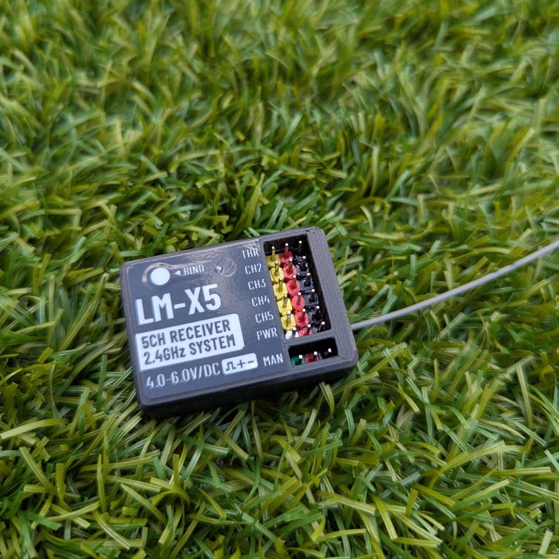 Puede incluir: Un receptor LM-X5 5CH negro, sistema de 2,4 GHz, con pines de colores y el texto "4.0-6.0V/DC". El dispositivo est&aacute; sobre hierba verde.