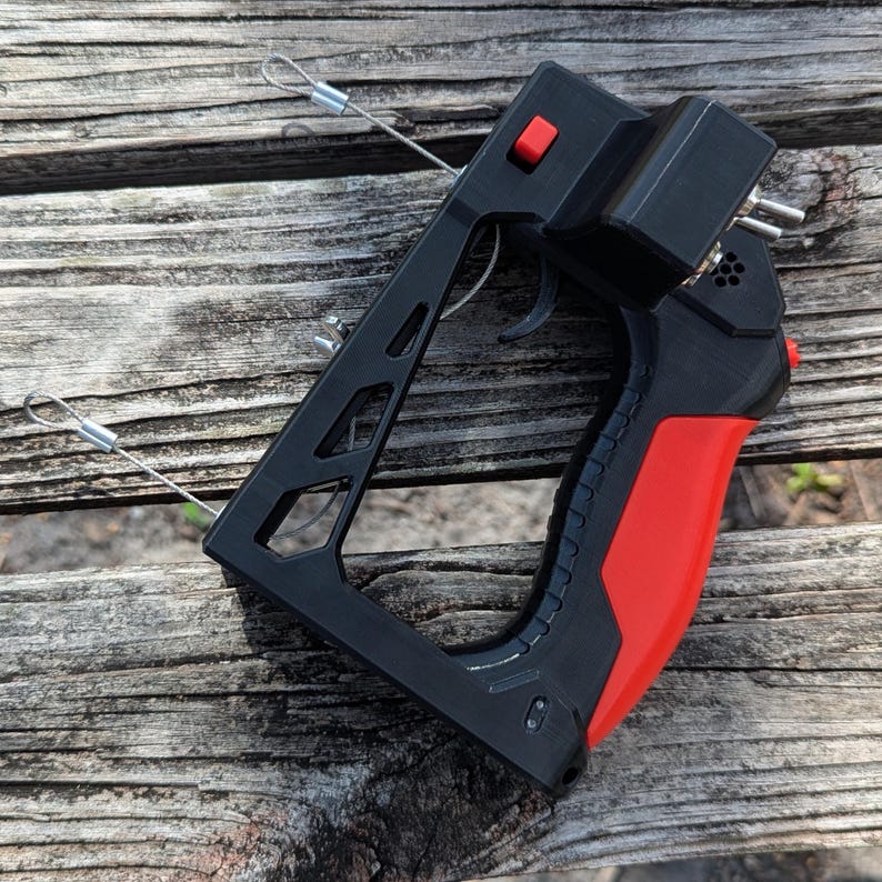 Puede incluir: Mando a distancia negro y rojo con gatillo y bot&oacute;n rojo. El mando tiene un marco negro con empu&ntilde;adura roja y una caja negra en la parte superior. Dos cables plateados se extienden desde la parte superior del mando.