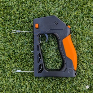 Puede incluir: Dispositivo port&aacute;til negro y naranja con gatillo y empu&ntilde;adura. El dispositivo tiene un cuerpo rectangular con una serie de agujeros y un mango curvo. Dos cables plateados se extienden desde el lateral. El dispositivo est&aacute; sobre un fondo de hierba verde.