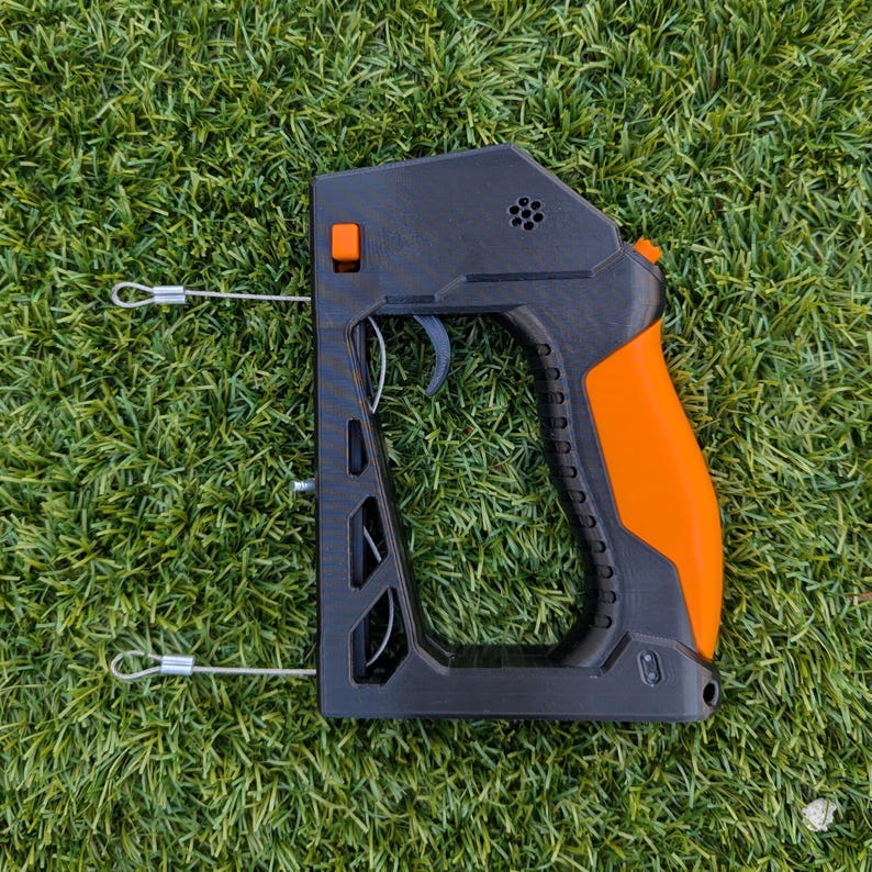 Puede incluir: Dispositivo port&aacute;til negro y naranja con gatillo y empu&ntilde;adura. El dispositivo tiene un cuerpo rectangular con una serie de agujeros y un mango curvo. Dos cables plateados se extienden desde el lateral. El dispositivo est&aacute; sobre un fondo de hierba verde.