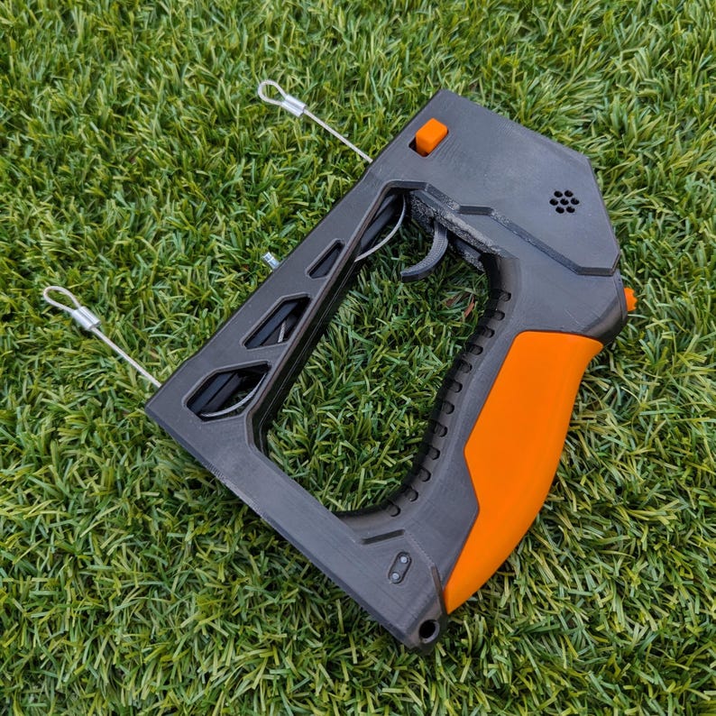 Puede incluir: Un dispositivo port&aacute;til negro y naranja con gatillo y mango. El dispositivo tiene un agarre texturizado y varios botones naranjas. Dos cables plateados se extienden desde la parte superior del dispositivo.