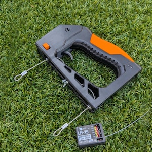 Puede incluir: Un dispositivo port&aacute;til negro y naranja con un gatillo y dos cables plateados adjuntos. Tambi&eacute;n hay un peque&ntilde;o dispositivo negro con el texto "LM-X5 2.4GHz System". Los art&iacute;culos est&aacute;n sobre un lecho de hierba verde.