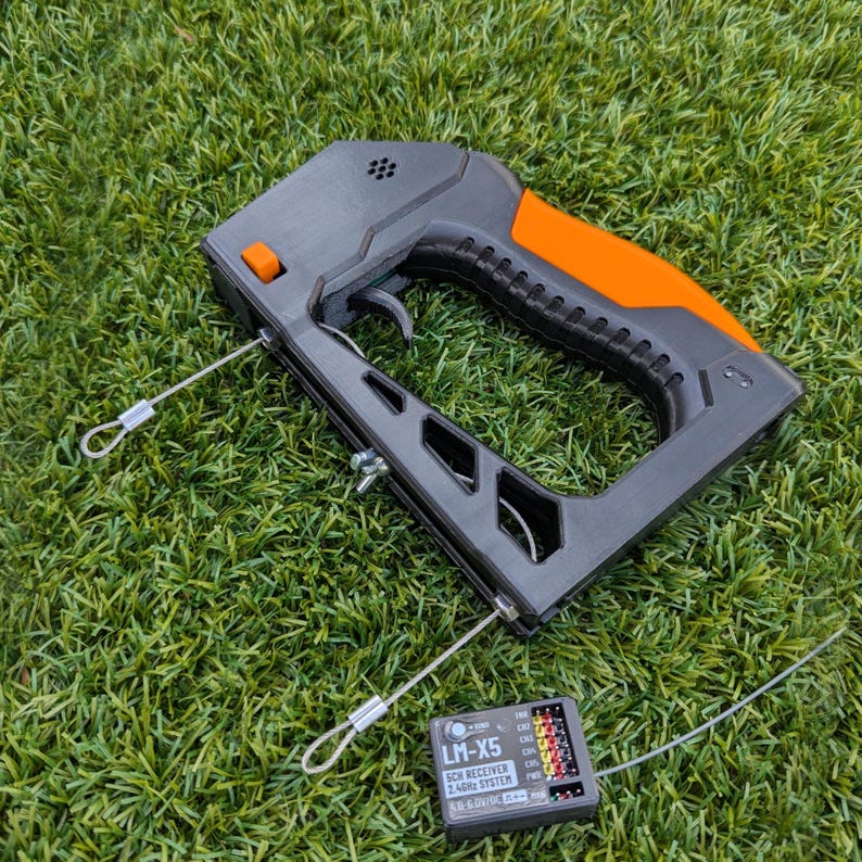 Puede incluir: Un dispositivo port&aacute;til negro y naranja con un gatillo y dos cables plateados adjuntos. Tambi&eacute;n hay un peque&ntilde;o dispositivo negro con el texto "LM-X5 2.4GHz System". Los art&iacute;culos est&aacute;n sobre un lecho de hierba verde.