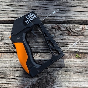 Puede incluir: Herramienta Zephyr Line Master negra y naranja con gatillo y dos cables met&aacute;licos adjuntos. El texto "Line Master Zephyr" est&aacute; impreso en el lateral. La herramienta descansa sobre una superficie de madera desgastada.