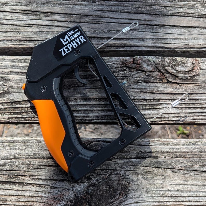 Puede incluir: Herramienta Zephyr Line Master negra y naranja con gatillo y dos cables met&aacute;licos adjuntos. El texto "Line Master Zephyr" est&aacute; impreso en el lateral. La herramienta descansa sobre una superficie de madera desgastada.