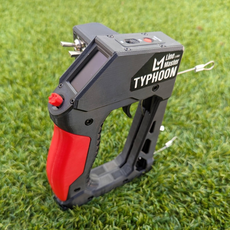 Puede incluir: Dispositivo Line Master Typhoon negro y rojo con empu&ntilde;adura de gatillo, pantalla peque&ntilde;a y componentes met&aacute;licos plateados. La palabra "TYPHOON" est&aacute; impresa en el lateral. El dispositivo est&aacute; sobre una superficie de hierba verde.
