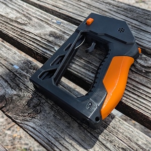 Puede incluir: Un dispositivo port&aacute;til negro y naranja con gatillo y empu&ntilde;adura. El dispositivo tiene un dise&ntilde;o angular con una empu&ntilde;adura naranja y un cuerpo negro. El gatillo es plateado. El dispositivo est&aacute; sobre una superficie de madera desgastada.