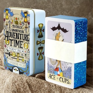 May include: A light blue metal tin and a deck of tarot cards. The tin features the words "A Totally Unofficial Adventure Time Tarot Deck" and illustrations. The deck is wrapped in white paper and has blue glitter edges. The Ace of Cups card is visible.