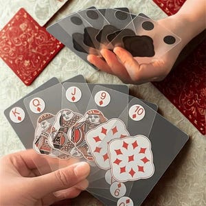 以下が含まれることがあります： ユニークなデザインのトランプ。カードは透明感があり、裏面はグレー、ダイヤのマークは赤色です。絵札には王族の詳細なイラストが描かれています。カードは人の手で持たれています。