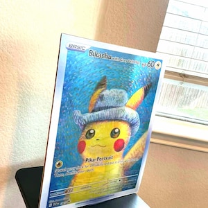 May include: A framed Pokémon trading card featuring Pikachu wearing a grey felt hat. The card has a blue background and the text "Sikachu with Grey Felt Hat" and "Pika-Portrait". The card is displayed on a black stand.