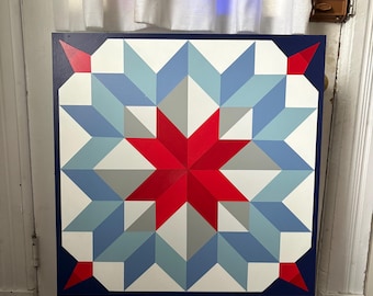 May include: A square, painted wooden wall hanging featuring a geometric star design. The artwork showcases a central red star surrounded by blue, gray, and white shapes, all framed by a dark blue border. The piece is displayed on a wooden easel.