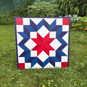 May include: Square wooden wall art featuring a star quilt design in red, white, and blue. The central star is red, surrounded by blue and white star shapes. The artwork is displayed outdoors on a grassy lawn.