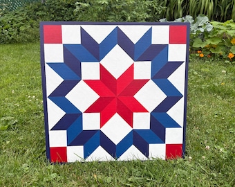 May include: Square wooden wall art featuring a star quilt design in red, white, and blue. The central star is red, surrounded by blue and white star shapes. The artwork is displayed outdoors on a grassy lawn.