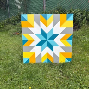 May include: A square, geometric quilt block painted with a starburst design. The star is composed of teal, white, and gray triangles, surrounded by yellow and orange triangles. The background is gray. The artwork is displayed on a grassy lawn.
