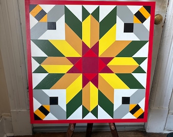 May include: A square, colorful quilt block design with a red border. The geometric pattern features a central starburst in red, yellow, and orange, surrounded by green, gray, and white shapes. Four stylized bees are in the corners.