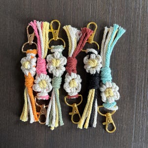 Puede incluir: Un conjunto de seis llaveros de macramé coloridos con diseños en forma de margarita y cierres de metal dorado. Cada llavero presenta una combinación de colores de hilo diferente, incluyendo naranja, rosa, verde, rojo, negro y azul.