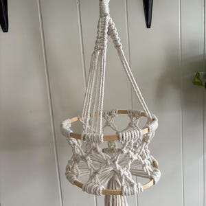 May include: An off-white macrame plant hanger. It features a wooden ring at the top for hanging, and wooden dowels incorporated into the design. The woven basket is designed to hold a small potted plant. The macrame is suspended from a wooden ring.