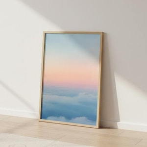 May include: A framed art print featuring a serene landscape of clouds and sky. The artwork displays a gradient of colors, transitioning from blue at the bottom to pink and orange in the middle, and light blue at the top. The frame is made of light-colored wood.
